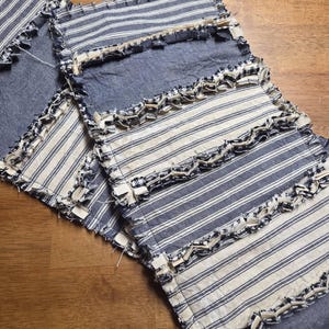 May include: A rectangular fabric runner featuring alternating panels of blue and white striped fabric and solid blue fabric. The edges of each panel are finished with a frayed, raw-edge seam detail that creates a textured, layered appearance.