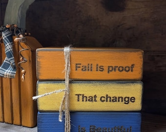 May include: A stack of three rectangular wooden blocks in orange, yellow, and blue, tied together with twine. The orange block reads Fall Is proof, the yellow block reads That change, and the blue block reads Is Beautiful in black text.