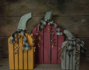 May include: Three decorative wooden pumpkins in yellow, deep red, and gray. Each pumpkin features a curved stem, a plaid fabric ribbon tied around the neck, and decorative brown wire tendrils with small wooden beads hanging down the front.