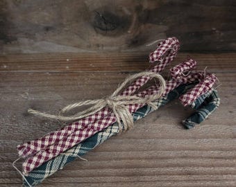 May include: A bundle of three fabric candy cane ornaments tied together with a piece of natural jute twine. Two of the candy canes feature a red and white gingham check pattern, while the third candy cane has a dark green and white plaid pattern.
