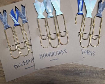 May include: Gold-toned paperclip bookmarks with decorative ribbon accents in blue, silver, and white. The bookmarks are attached to brown cardstock with the word "BOOKMARKS" printed on them. A simple and elegant design.