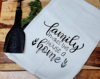 May include: White kitchen towel with black text that reads "family makes this house a home" with a small leaf design on either side of the text.