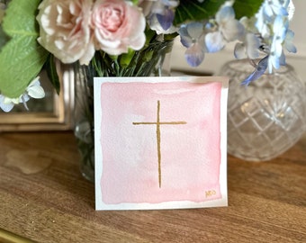May include: A watercolor painting of a gold cross on a pink background. The painting is signed in the bottom right corner.