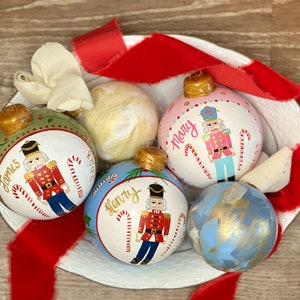 Puede incluir: Una colección de adornos navideños pintados a mano en un cuenco blanco, acentuados con una cinta roja. Los adornos presentan diseños de cascanueces y nombres personalizados como "James", "Mary" y "Henry". Cada adorno tiene una tapa dorada y está decorado con bastones de caramelo.