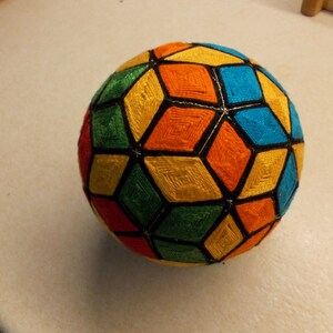 May include: A colorful, geometric ball made of yarn with a black outline. The ball is made of a variety of colors, including yellow, orange, red, green, and blue.