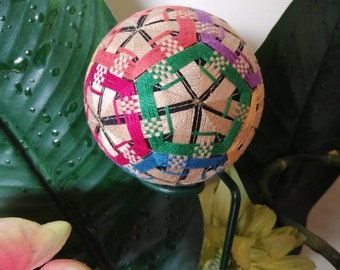May include: A colourful, hand-stitched Japanese ball known as a "Temari" with a geometric pattern in shades of pink, green, blue, and purple. The ball is made of fabric and thread and is displayed on a green stem.