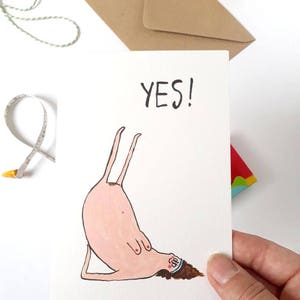 May include: A white card with a hand-drawn illustration of a person doing a yoga pose. The person is pink with brown hair and is wearing a blue headband. The text "YES!" is written in black at the top of the card.