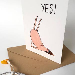 May include: A white card with a pink cartoon figure doing a yoga pose with the word "YES!" written in black above it.