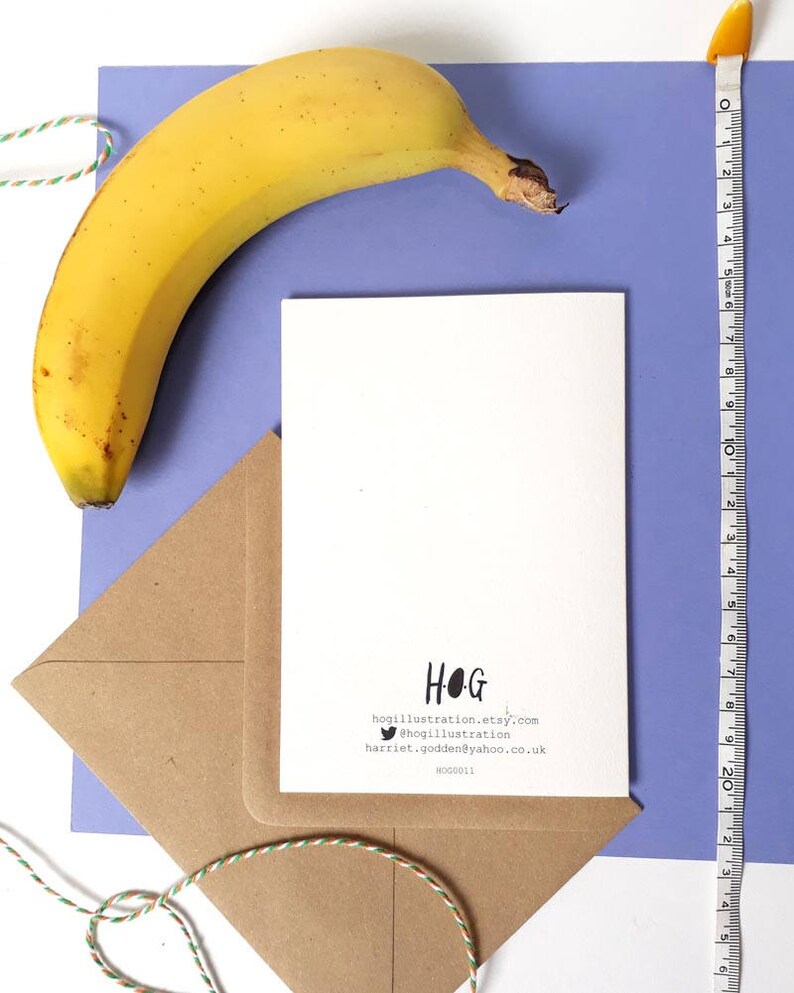 May include: A white card with black text that reads "HOG" and includes contact information for the artist. The card is on a brown envelope with a blue background. A yellow banana is in the upper left corner of the image.