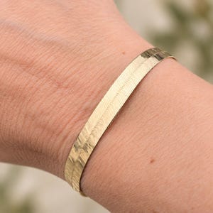 May include: A gold-toned herringbone chain bracelet featuring a flat, flexible design with a repeating V-shaped pattern. The metallic surface has a polished finish that reflects light, creating a smooth, continuous band around the wrist area.