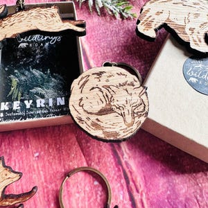 May include: Wooden keychains with animal designs, including a sleeping fox, a running hare, and a bear. The keychains are light brown and appear to be made of wood. The keychains are displayed with small boxes and a sprig of greenery.