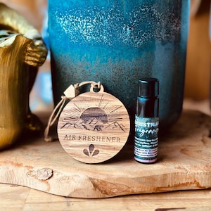 Könnte beinhalten: Ein Holz-Lufterfrischer mit einer Bergszene und den Worten "Air Freshener" eingraviert. Daneben befindet sich eine kleine Flasche Weihnachtsduftöl.