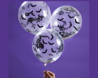 May include: Three clear balloons with black bat confetti inside. The balloons are tied together with a string and held by a hand.