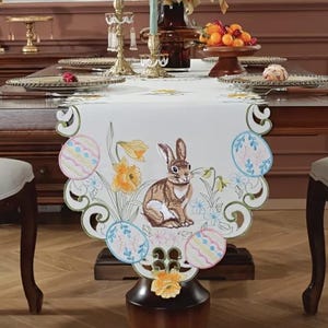 以下が含まれることがあります： 白い刺繍のテーブルランナーに、茶色のウサギ、カラフルなイースターエッグ、黄色の花が描かれています。ランナーは木製のテーブルに置かれ、白い椅子が置かれています。