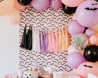 May include: A pink and purple balloon arch with black bats, a black and white bat patterned backdrop, and a table set with pink and white plates and cups for a Halloween party.