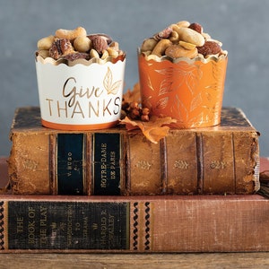 Op de afbeelding: Twee papieren bekers gevuld met gemengde noten, &eacute;&eacute;n wit met "Give Thanks" in gouden letters en een bladmotief, de andere oranje met een bladpatroon. Ze staan op oude boeken, wat wijst op een herfstthema.