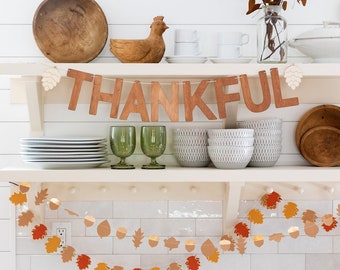 Op de afbeelding: Een keukenkastje met herfstdecoratie. Een houten banner met de tekst "THANKFUL" hangt boven een slinger van papieren bladeren en eikels. Andere items zijn houten kommen, witte borden, groene glazen en een houten kippenornament.