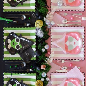 May include: Halloween party table setting with green and white striped plates, black witch-shaped plates, and pink plates. The witch plates have a green face and black hat. Silverware, napkins, and decorative elements complete the festive arrangement.