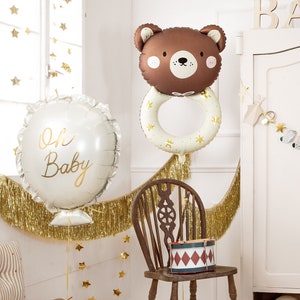 May include: A baby shower party decoration set with a brown bear balloon, a white balloon with the text "Oh Baby", gold fringe, and a wooden sign that says "BABY".