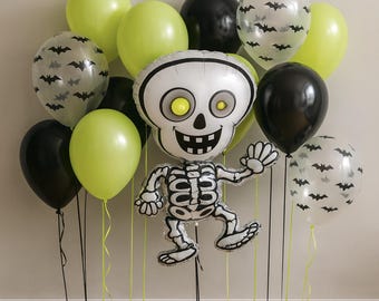 May include: A festive Halloween balloon arrangement featuring a large skeleton-shaped foil balloon with yellow eyes, surrounded by black, lime green, and bat-printed balloons. The balloons are tied with black and yellow ribbons.
