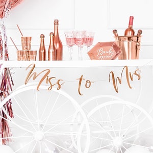 Könnte beinhalten: Ein weißer Barwagen mit roségoldenen Akzenten, dekoriert für eine Bridal Shower. Der Wagen ist mit roségoldenen Luftschlangen, einem roségoldenen "Miss to Mrs" Banner und einem roségoldenen Eiseimer mit Weinflaschen geschmückt. Es gibt auch ein roségoldenes Schild mit der Aufschrift "Bride Squad".