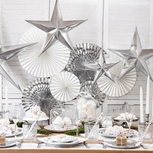 May include: A white table setting with silver accents. The table is decorated with white paper fans, silver stars, and white marshmallows in glass jars. The place setting includes a white plate, a silver charger, a clear glass, and a silver napkin ring. The table is set for a special occasion, such as a wedding or a holiday party.