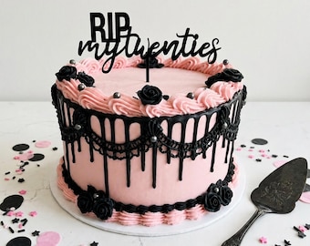 May include: A round cake with light pink frosting, black icing drips, and black piped roses. A black cake topper reads RIP my twenties. The cake is decorated with small silver spheres and sits alongside a metal cake server on a white surface.