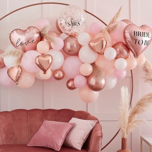 Puede incluir: Un arco de globos de color oro rosa con globos rosas, melocotón y blancos. El arco está decorado con hierba de la pampa e incluye globos con el texto "love" y "bride to be".
