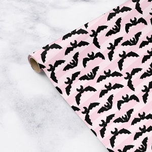 Op de afbeelding: Roze en zwart inpakpapier met een herhalend patroon van vleermuizen. De vleermuizen zijn zwart en hebben een eenvoudig ontwerp. De achtergrond is roze met witte strepen.