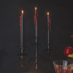 May include: A black metal candelabra holding three black taper candles with red wax dripping down the sides. The candles are lit, casting a warm glow. To the right, a tiered serving stand displays various fruits, cookies, and small treats.