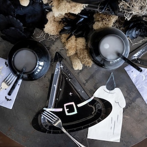 May include: A black witch hat-shaped paper plate with a holographic buckle design, accompanied by white napkins featuring moon, star, and bat patterns, silver forks, black cups with straws, and a large black cauldron surrounded by beige moss.
