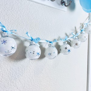 May include: A garland of clear, round ornaments with snowflake designs and blue ribbons. The ornaments are strung on a white string and hang against a white wall. Balloons in shades of blue and purple are visible in the background.