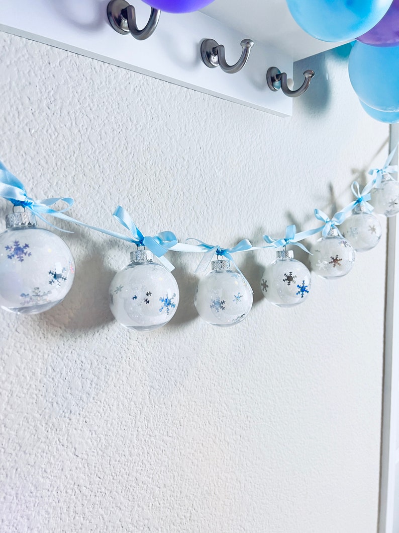 May include: A garland of clear, round ornaments with snowflake designs and blue ribbons. The ornaments are strung on a white string and hang against a white wall. Balloons in shades of blue and purple are visible in the background.