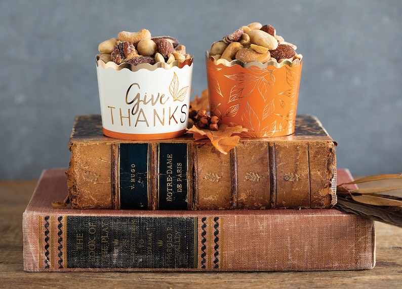 Op de afbeelding: Twee papieren bekers gevuld met gemengde noten, &eacute;&eacute;n wit met "Give Thanks" in gouden letters en een bladmotief, de andere oranje met een bladpatroon. Ze staan op oude boeken, wat wijst op een herfstthema.