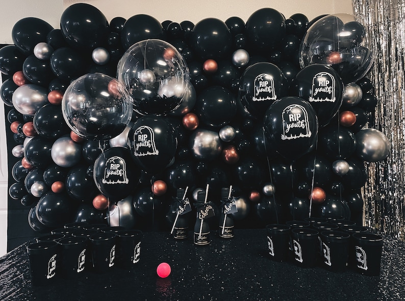 May include: A black and silver balloon arch with the text "RIP youth" on several balloons. There are also black and silver cups in front of the arch.