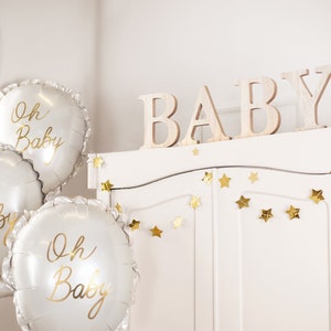 May include: Three white balloons with gold lettering that reads "Oh Baby" are in front of a white cabinet with gold star garland. The cabinet has the word "BABY" spelled out in wooden letters on top.