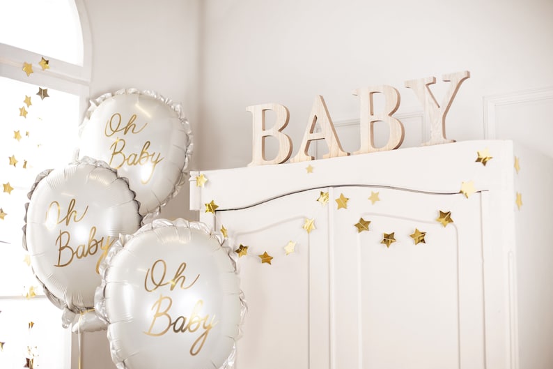 May include: Three white balloons with gold lettering that reads "Oh Baby" are in front of a white cabinet with gold star garland. The cabinet has the word "BABY" spelled out in wooden letters on top.
