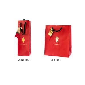 May include: Two red gift bags with gold detailing. One is a wine bag with a gold lightbulb graphic and the text "GETTING LIT". The other is a gift bag with a gingerbread man graphic and the text "OH SNAP". Both have black handles.