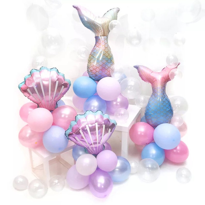 May include: Three balloon centerpieces featuring mermaid tails and seashells. Each centerpiece is made of pastel pink, blue, and purple balloons with clear balloons. The mermaid tails are iridescent and have a scale pattern. The seashells are iridescent and have a pearl-like finish.