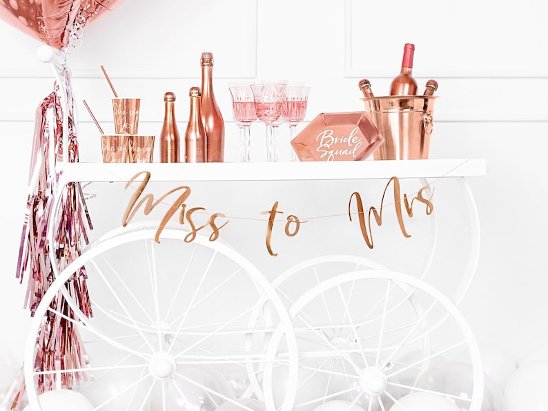 May include: A white bar cart with rose gold accents, decorated for a bridal shower. The cart is decorated with rose gold streamers, a rose gold "Miss to Mrs" banner, and a rose gold ice bucket with bottles of wine. There is also a rose gold sign that says "Bride Squad".