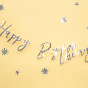 May include: Silver foil Happy Birthday banner with silver star confetti on a yellow background.