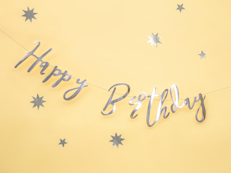 May include: Silver foil Happy Birthday banner with silver star confetti on a yellow background.