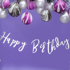 May include: Silver and purple balloons with a silver "Happy Birthday" banner hanging in front of a silver and purple iridescent background.