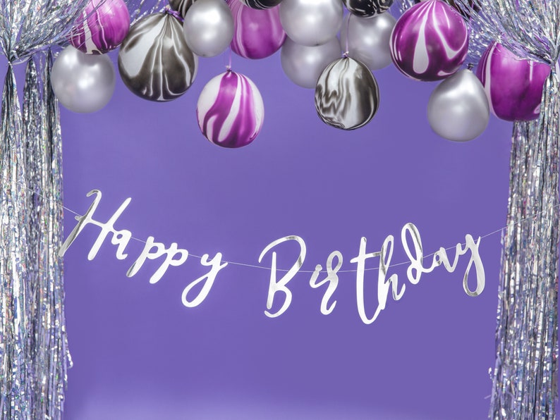 May include: Silver and purple balloons with a silver "Happy Birthday" banner hanging in front of a silver and purple iridescent background.