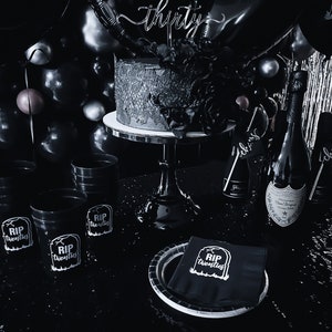 May include: A monochrome party scene featuring a cake with a 'thirty' topper, black balloons, and champagne. The cake is decorated with floral accents. Black cups and napkins display 'RIP twenties' text, creating a themed atmosphere.