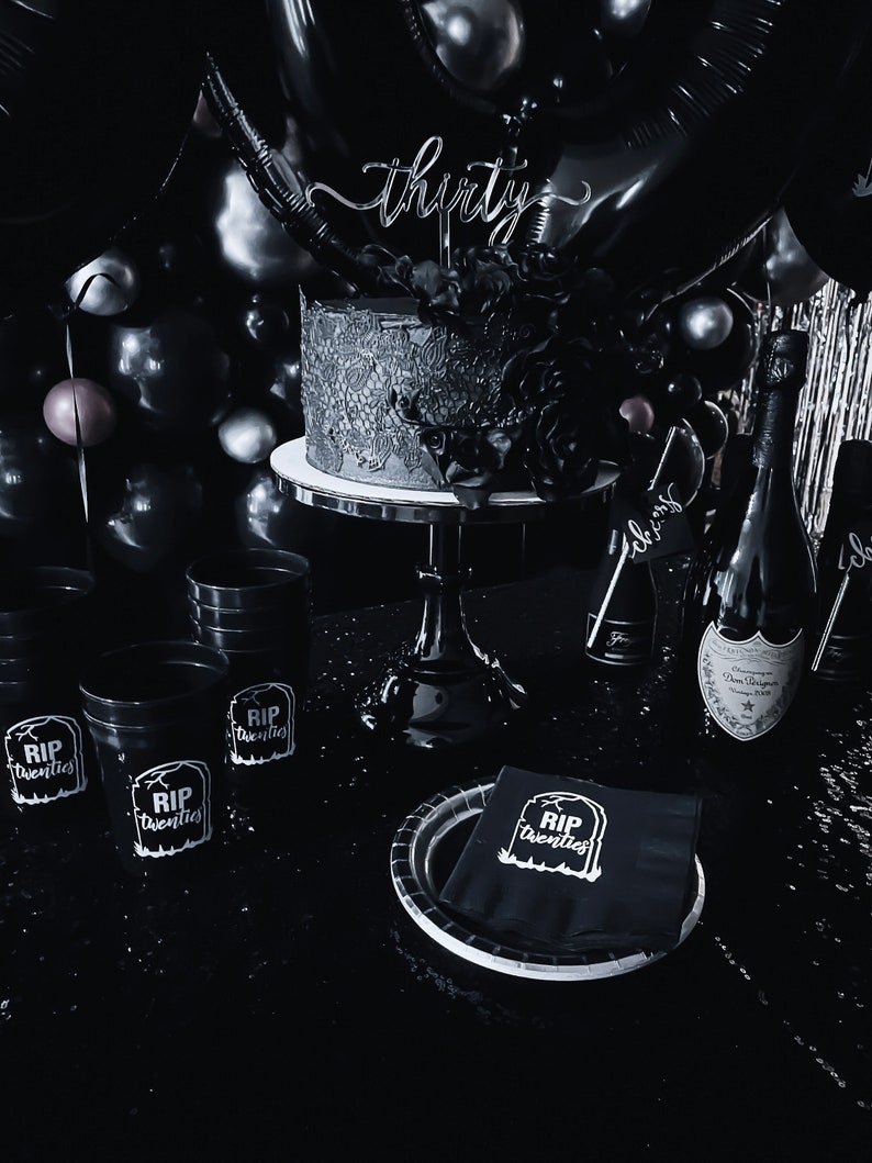 May include: A monochrome party scene featuring a cake with a 'thirty' topper, black balloons, and champagne. The cake is decorated with floral accents. Black cups and napkins display 'RIP twenties' text, creating a themed atmosphere.