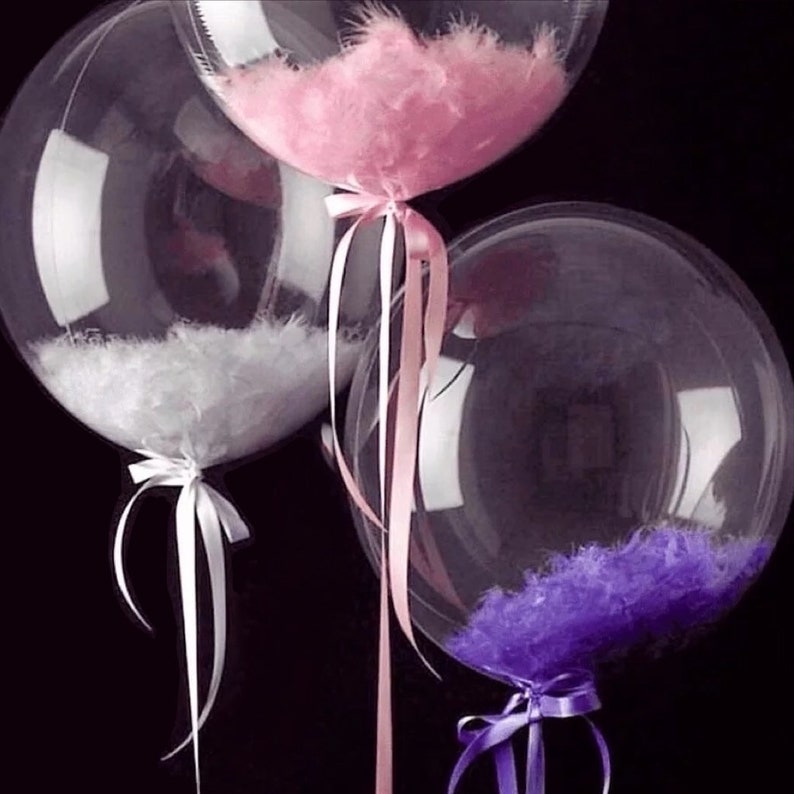 May include: Three clear plastic balloons filled with feathers. The balloons are tied with white, pink, and purple ribbons.