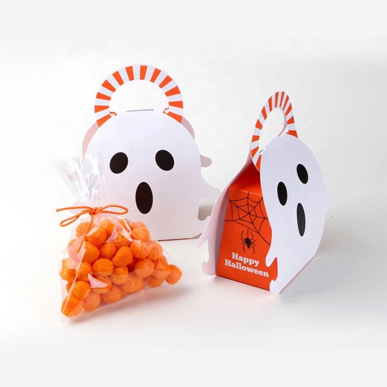 May include: Two white ghost-shaped paper treat boxes with orange and white striped handles. One box is open, revealing an orange side panel with a black spider web and spider graphic and the text Happy Halloween. A clear plastic bag of orange snacks.