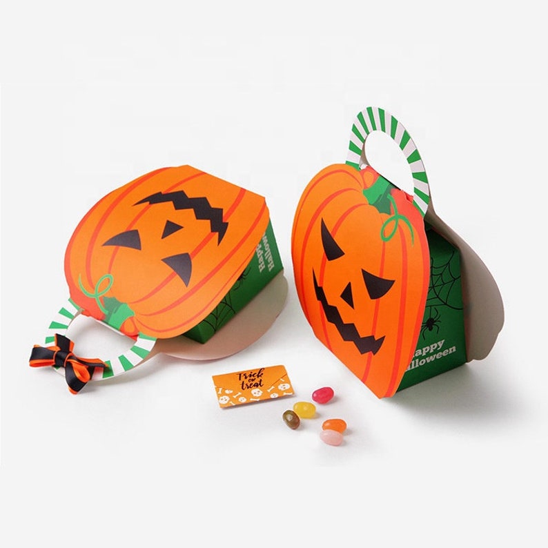 May include: Two cardboard treat boxes shaped like orange jack-o'-lanterns with green sides featuring spider web patterns and the text Happy Halloween. One box has a black and orange ribbon on the handle. A small card reads Trick or treat.