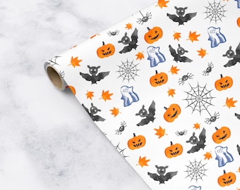 Puede incluir: Papel de regalo blanco con un patrón repetido de calabazas naranjas, murciélagos negros, telarañas grises y fantasmas grises. El papel está enrollado y el extremo es visible.